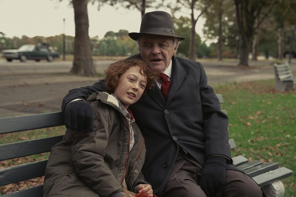 Girl with short red hair, fair skin, dark jacket with red details hugged by an old man, brown hat, white hair, fair skin, dark suit, red tie, black gloves.  The two are sitting on a bench in a park - Metropolis